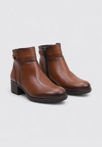 Botas de cuero marrón hasta el tobillo con punta redondeada, cremallera lateral y detalle de hebilla decorativa. Tacón bajo y textura suave.