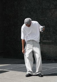 Sive široke sivo hlače, bela oversize majica, siva kapica, črne čevlje, drži kozarček za kavo in telefon, stoji na teksturirani površini.