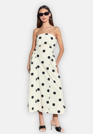 Frau trägt ein trägerloses weißes Midikleid mit großen schwarzen Punkten, schwarze hochhackige Sandalen und schwarze Sonnenbrille.