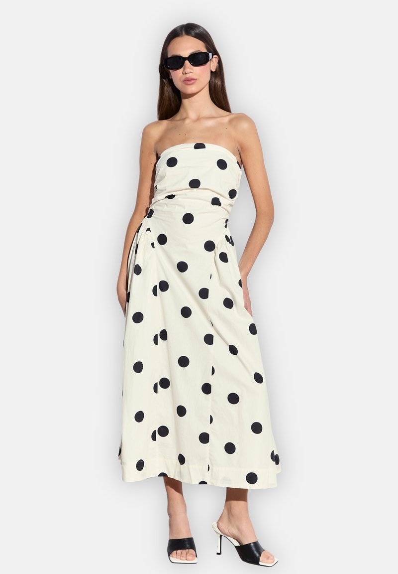 Frau trägt ein trägerloses weißes Midikleid mit großen schwarzen Punkten, schwarze hochhackige Sandalen und schwarze Sonnenbrille.