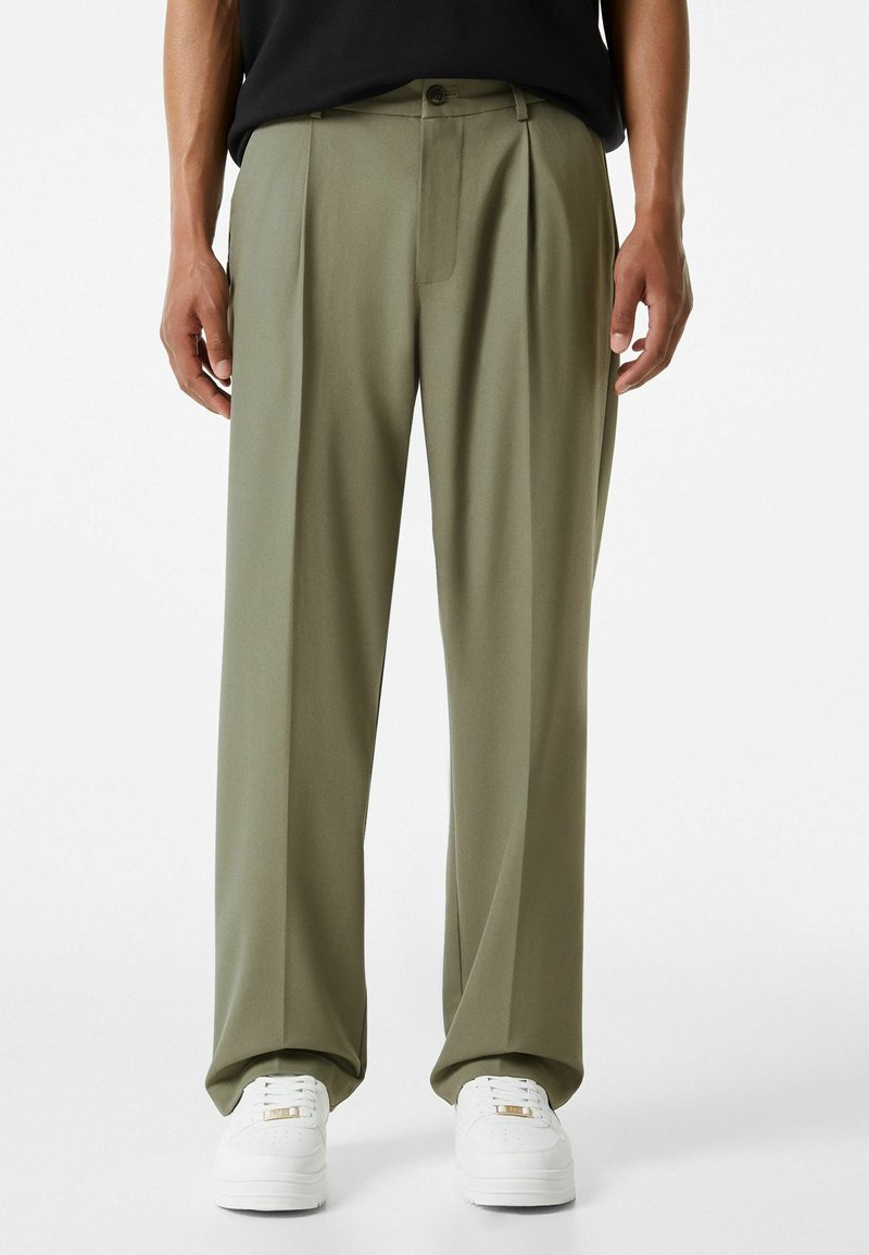 Olivově zelené plisované kalhoty vyrobené z hladkého materiálu, s přední kapsou, poutky na pásek a knoflíkovým zapínáním, spárované s bílými teniskami.