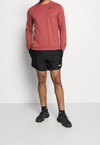 Chemise de sport rouge à manches longues avec logo Nike noir, associée à un short noir avec logo Nike blanc, et des baskets noires.