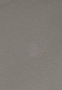 Grå ribbet skjorte med en glat tekstur, der har et lille, metallisk logo nederst til højre. Enkel, minimalistisk design.