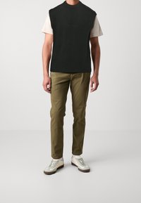 Black sleeveless knit sweater over a white t-shirt, paired with olive green trousers and cream sneakers. Flat texture, ribbed hem.