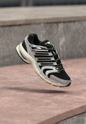 Czarno-srebrny but sportowy Adidas z siateczki i materiałów syntetycznych, unoszący się nad różowawą betonową powierzchnią i szarym kamiennym tłem.