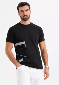 Černé bavlněné tričko s kulatým výstřihem a krátkými rukávy. Na spodní levé straně má šedý grafický design. Hladká textura.