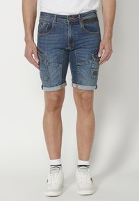 Denim šorti ar izbalējušu zilu tonu; iekļauj vairākas kabatas un sarullētas apakšmalas. Kombinēti ar baltām sporta apaviem un zeķēm.