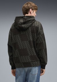 Felpa nera con cappuccio e motivo geometrico in verde scuro e grigio, con coulisse al cappuccio e polsini a coste. Indossata con jeans azzurri.