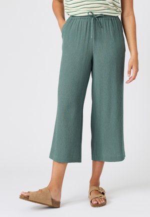 Frau trägt türkisfarbene, weit geschnittene, kurze Hosen mit elastischem Bund und Kordelzug, beige offene Sandalen, steht mit einer Hand in der Tasche.