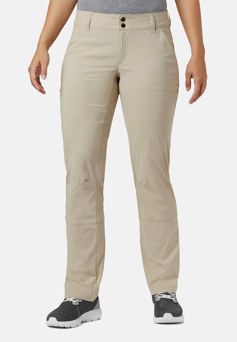 Beige Outdoor-Hosen mit schmaler Passform, zwei Fronttaschen, Doppelknebelverschluss und weichem, leichtem Stoff. Kombiniert mit grauen Sportschuhen.