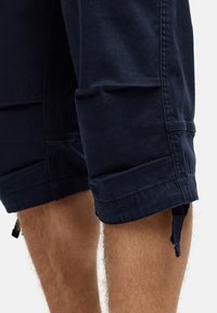 Navy blue cotton shorts with rolled hems, featuring side pockets and adjustable drawstrings at the cuffs. Smooth texture, casual design.