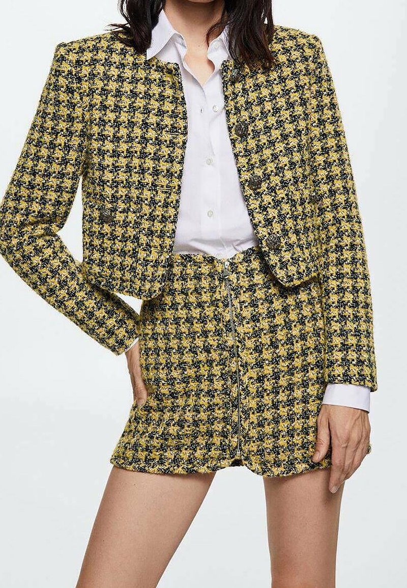 Femme portant une veste courte en tweed pied-de-poule jaune et noire et une mini-jupe taille haute assortie, associées à une chemise blanche boutonnée.