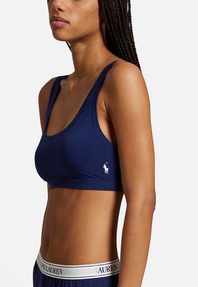 Sportovní podprsenka tmavě modré barvy z měkké látky s širokými ramínky a vypasovaným střihem. Na levé straně je malý bílý logo.