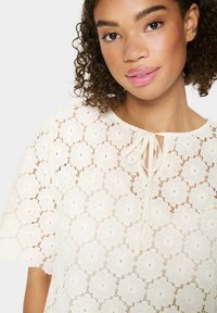 Top bianco in pizzo con motivi floreali, maniche corte e scollatura con un nastro annodato. Tessuto delicato e strutturato.