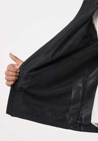 Hånd trækker en sort jakke op, der viser sort mesh-foring og lynlåsdetalje på ens hvid baggrund.