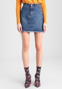 Vrouw met een blauwe denim rok met hoge taille en rafelige zoom, een mosterdkleurige geribbelde trui en zwarte enkellaarsjes met bloemmotief.