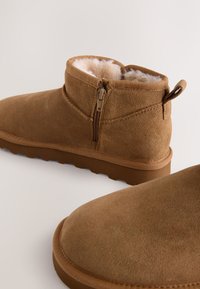 Bottines en daim marron avec fermeture éclair latérale et doublure en fourrure blanche douce, présentées sur un fond clair.