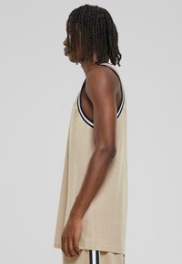Beige nettopp med løs passform, med svart kant langs halsutringningen og armhullene. Glatt tekstur, ermeløs design.