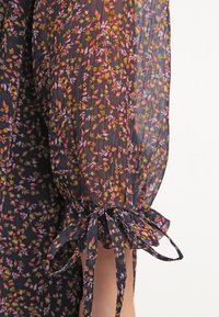 Manche en tissu noir transparent à motif floral rose et jaune, froncée et attachée au poignet avec un nœud assorti en tissu.