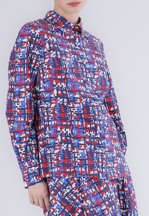 Persoon draagt een shirt met lange mouwen en een rok met een abstract geometrisch patroon in rood, blauw, wit en zwart tegen een effen achtergrond.