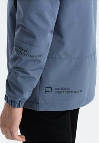 Giacca prestazionale grigia con finitura testurizzata. Presenta polsini elastici e logo stampato sulla manica. Materiale leggero e traspirante.