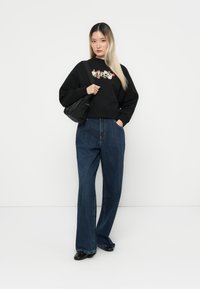 Felpa nera con ricami floreali, abbinata a jeans a gamba larga blu scuro. Il modello tiene una borsa nera e si trova di fronte a uno sfondo bianco.