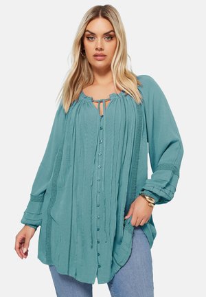 Teal farbige Bluse mit Knopfverschluss, plissiertem Stoff, Kordelzug am Hals und Spitzelementen. Sie hat lange Ärmel und einen lässigen Schnitt, kombiniert mit einer blauen Jeans.