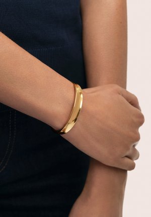 Brazalete de oro en forma de pulsera, llevado en la muñeca, con una superficie lisa y pulida. El diseño es elegante con una forma circular sencilla.