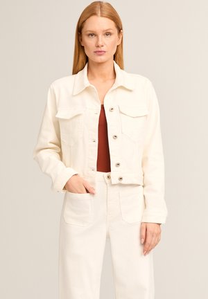 Frau mit glattem, hellbraunem Haar trägt eine weiße, geknöpfte Jacke und passende hoch taillierte Hosen mit Taschen über einem rostfarbenen Oberteil.
