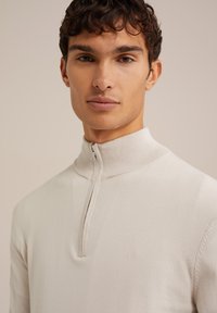 Svetlo bež pulover s visokim ovratnikom in pol zadrgo. Narejen je iz gladkega pletenega materiala; prilega se in prikazuje čist silhuet.