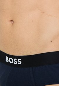 Mørkeblå bomulls truse med svart elastisk midje som har hvit "BOSS" tekst. Nærbilde av nedre torso som viser tekstur og hud.