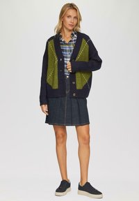 Marineblauer Cardigan mit grünen Rautenmuster, strukturiertem Strick, Knopfverschluss, über einem karierten Hemd getragen, kombiniert mit einem denim Faltenrock und schwarzen Sneakers.