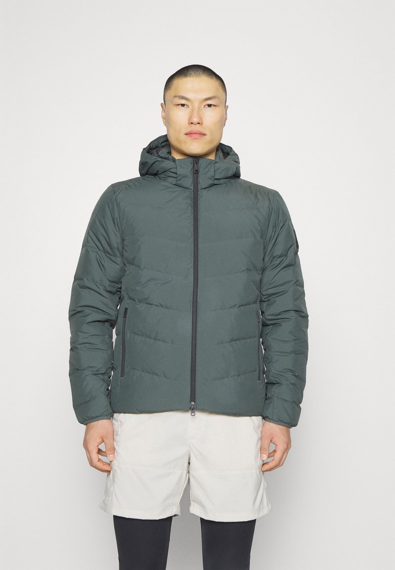 Plüschige grüne Jacke mit Kapuze, vorderem Reißverschluss und Seitentaschen. Hat ein gestepptes Muster und eine weiche Textur. Wird über Shorts getragen.