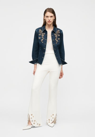 Γυναίκα που φορά σκούρο τζιν μπουφάν με λουλουδάτες διακοσμήσεις, λευκό διαφανές τοπ, φαρδιά λευκά παντελόνια με λουλουδάτες λεπτομέρειες και λευκά μυτερά παπούτσια.