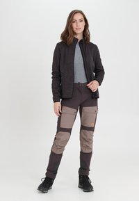 Zwarte zip-up jas over een grijs shirt, gecombineerd met donkere, meer-pocket broeken met lichtere stoffen accenten. Zwarte schoenen complementeren de outfit.