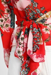 Blusa floreale rossa con cintura annodata, tessuto trasparente e motivi floreali colorati in rosa e blu su uno sfondo rosso brillante.