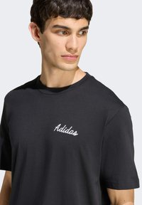Black cotton t-shirt with short sleeves, featuring white embroidered "Adidas" logo on the upper left chest area. Smooth texture, relaxed fit.