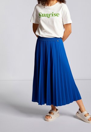 Falda azul plisada con cintura elástica, combinada con una camiseta blanca con texto verde. El conjunto se completa con sandalias de suela gruesa.