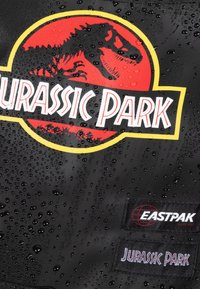 Mochila de tela negra con un logo de Jurassic Park en rojo y amarillo que incluye la silueta de un dinosaurio, gotas de agua y etiquetas de la marca Eastpak.
