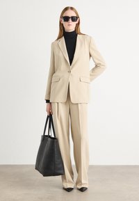 Beige tailored suit with a single-button blazer and wide-leg trousers, paired with a black turtleneck and large black tote bag.