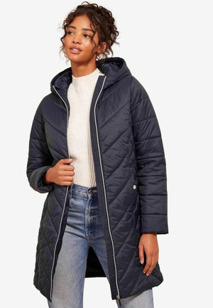 Marineblauwe gevoerde lange jas met capuchon, voorzien van diamantpatronen, een ritssluiting aan de voorkant en zijzakken, gedragen over een lichte trui en jeans.