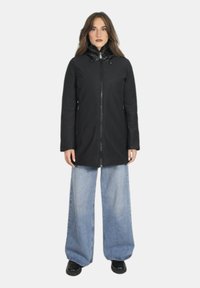 Cappotto nero con chiusura a zip frontale, colletto alto e cordoncino. Indossato sopra jeans a gamba larga blu chiaro e scarpe nere. Texture liscia.