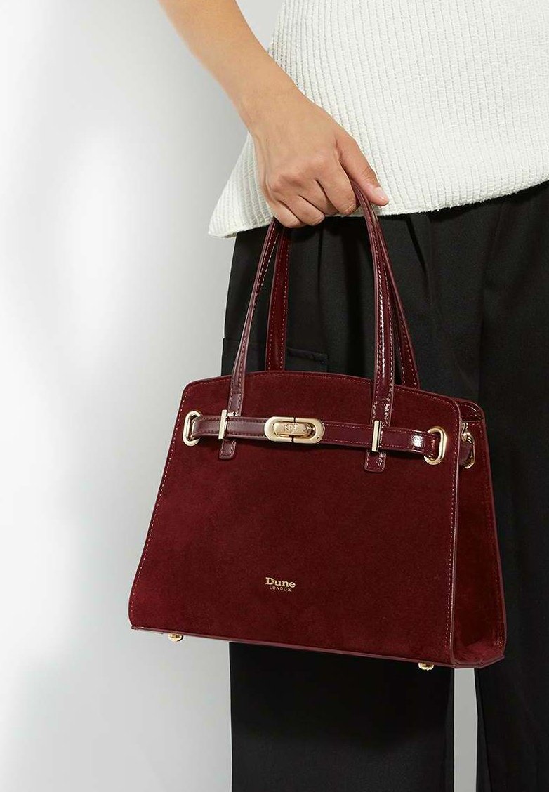 Burgundy suede handbag with gold hardware, structured design, short handles, and a logo at the base. Textured surface with shiny accents.