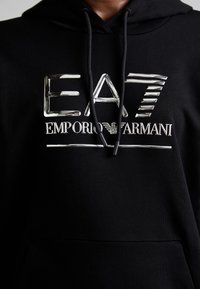 Svart hettegenser med en blank sølvfarge logo "EA7 EMPORIO ARMANI" over brystet, med en klassisk snorhette. Myk tekstur.