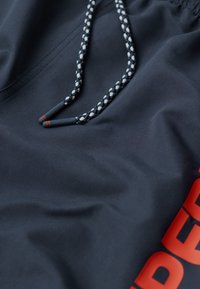 Pantalones cortos de tejido azul marino oscuro con una textura suave, que cuentan con un cordón trenzado y una gran impresión de logotipo rojo en el costado.