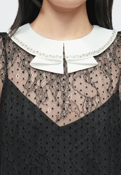 Tela de encaje negra con un cuello blanco que tiene detalles de brillantes. El cuello presenta un diseño de doble lengüeta y está superpuesto sobre una base transparente con puntos.