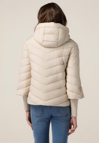 Beige pufferjakke med et chevronmønster, hætte og korte ærmer, lavet af tekstureret stof. Båret over blå jeans.