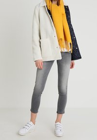 Gewaxt witte en marineblauwe doorgestikte jas, gele sjaal, grijze jeans en witte sneakers met klittenbandsluitingen. Casual outfit met contrasterende kleuren.