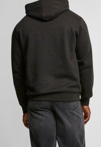 Černá mikina s kapucí prostornou kapucí, měkkou strukturou a pohodlným střihem. Kombinována s tmavě šedými džíny, které mají standardní zadní kapsy.