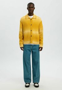 Vyras dėvi geltonos spalvos gradientinį megztinį su sagomis, baltą apatinį marškinėlį, laisvus mėlynus džinsus ir juodai baltus batus, stovintis priešais paprastą foną.